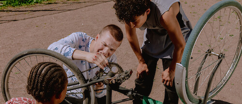 A young man with bloodied knuckles tries to fix a bicycle chain, his friends watching on.