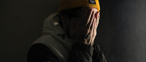 A man wearing a hoodie and yellow beanie, covering his face in despair